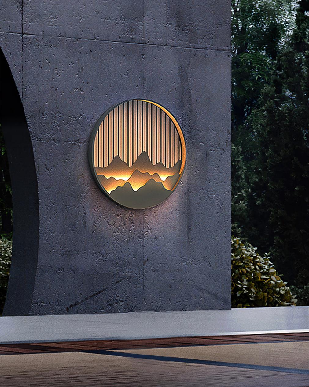 Lit mountain wall light on rough dark concrete wall above stone ledge, with green foliage at right