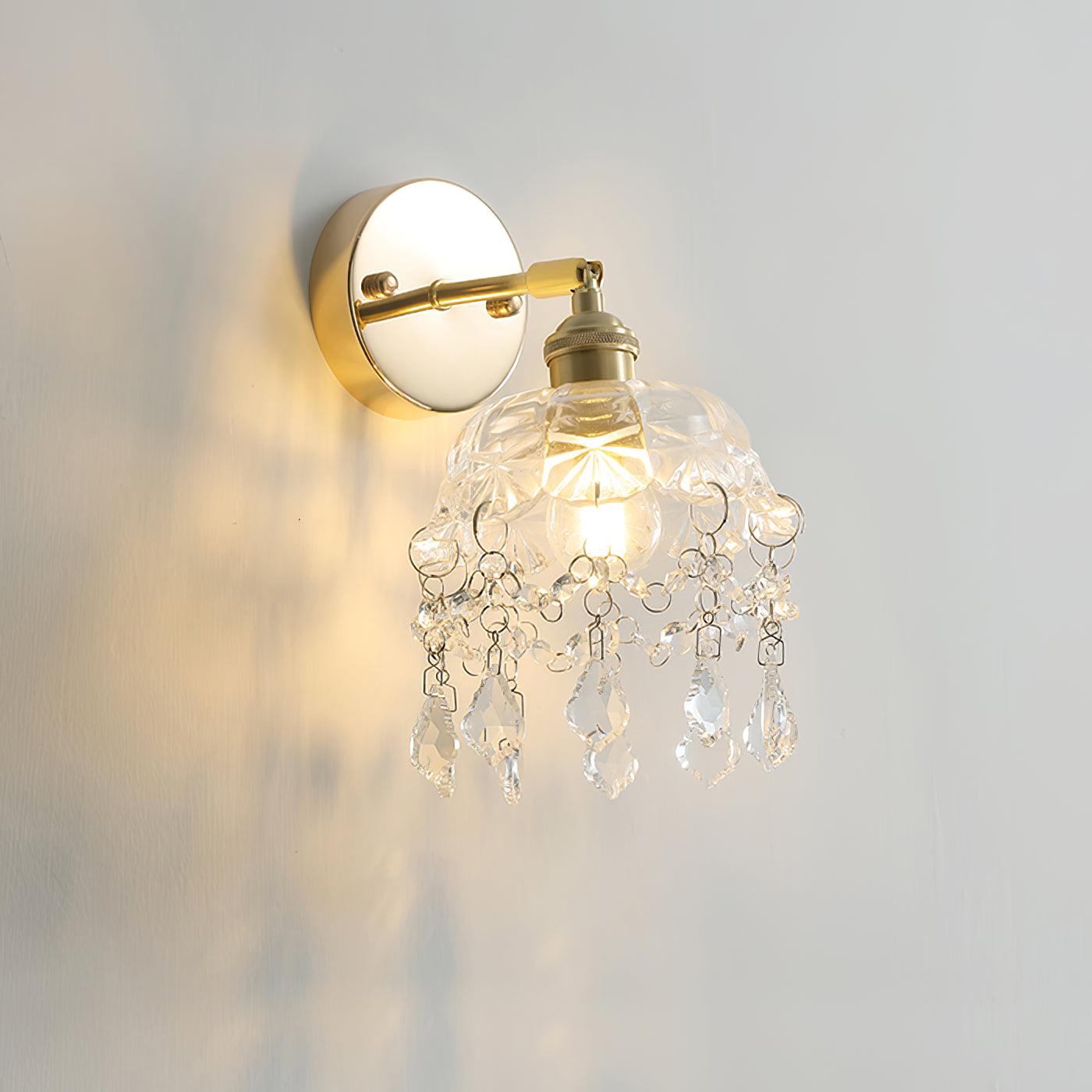 Lit clear bell and crystal drop fringe below a gold-tone arm and round wall plate on a pale wall