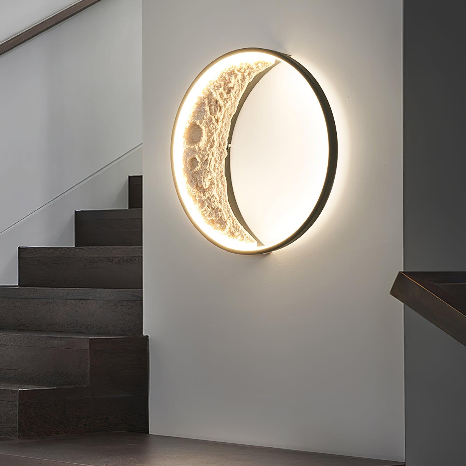 Illuminated crater-textured crescent in a black circular wall-light frame beside dark wooden stairs