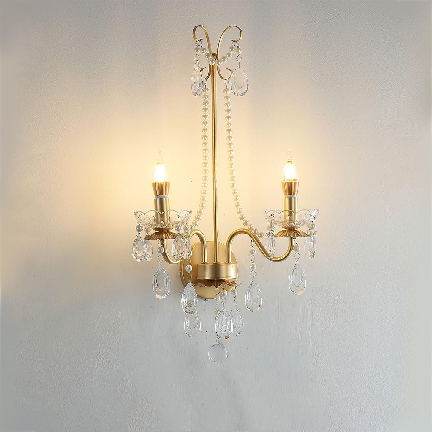 Lit front gold-and-clear fixture on textured wall, with two curved arms, long bead chains, teardrop-shaped drops and round rear plate