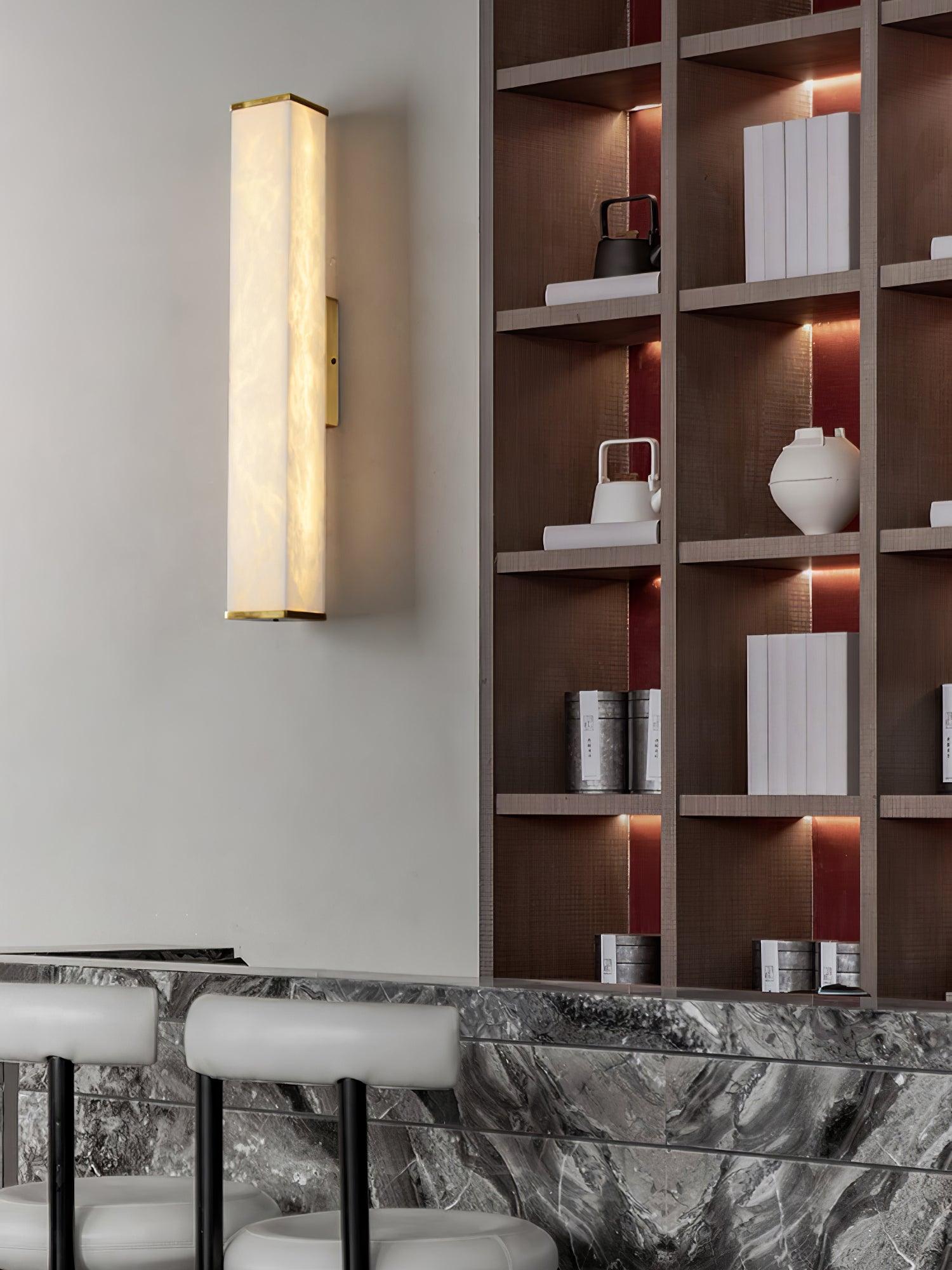 Lit alabaster wall light with brass trim beside wooden shelves holding books and teapots above a stone-pattern counter