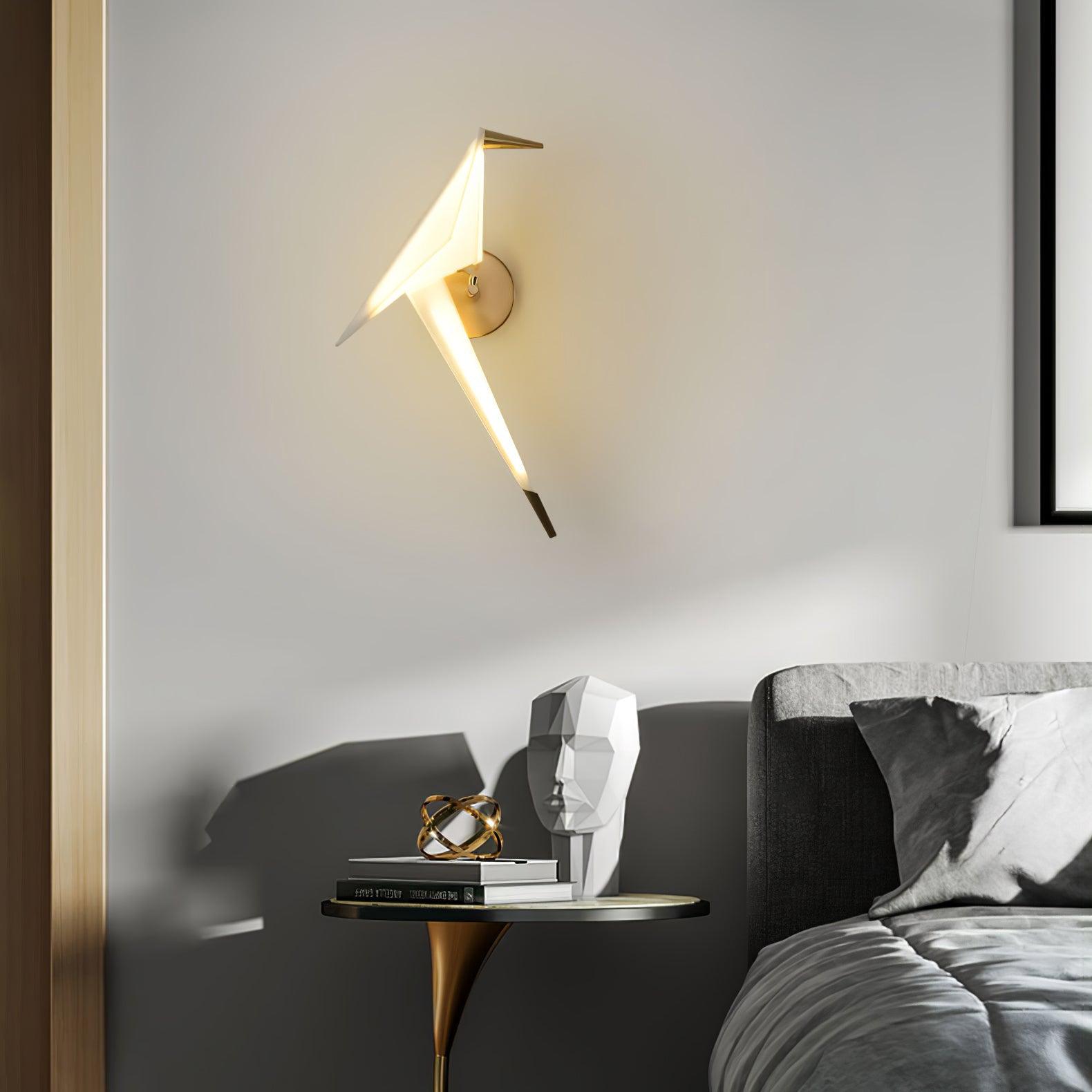 Gold and White bird wall light glowing beside a gray bed and round side table with books and a geometric head ornament