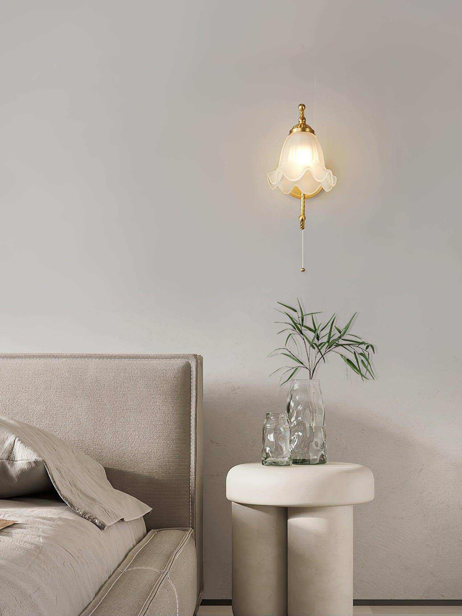 Front lit floral glass wall lamp above small white bedside table with glass vases, beside upholstered bedhead