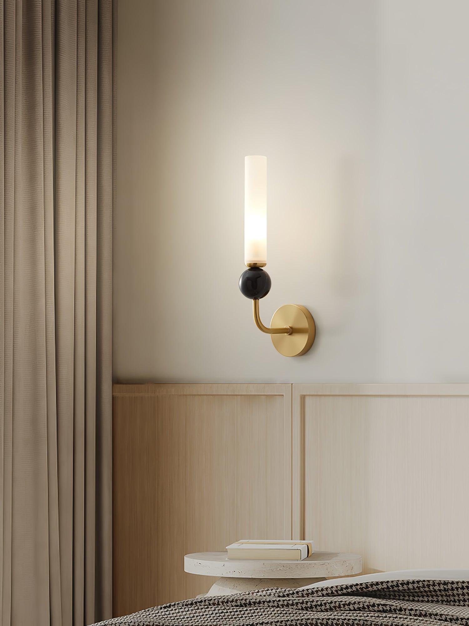 Curved brass wall light with marble sphere and lit white cylinder beside bed, above stone table and books near curtain