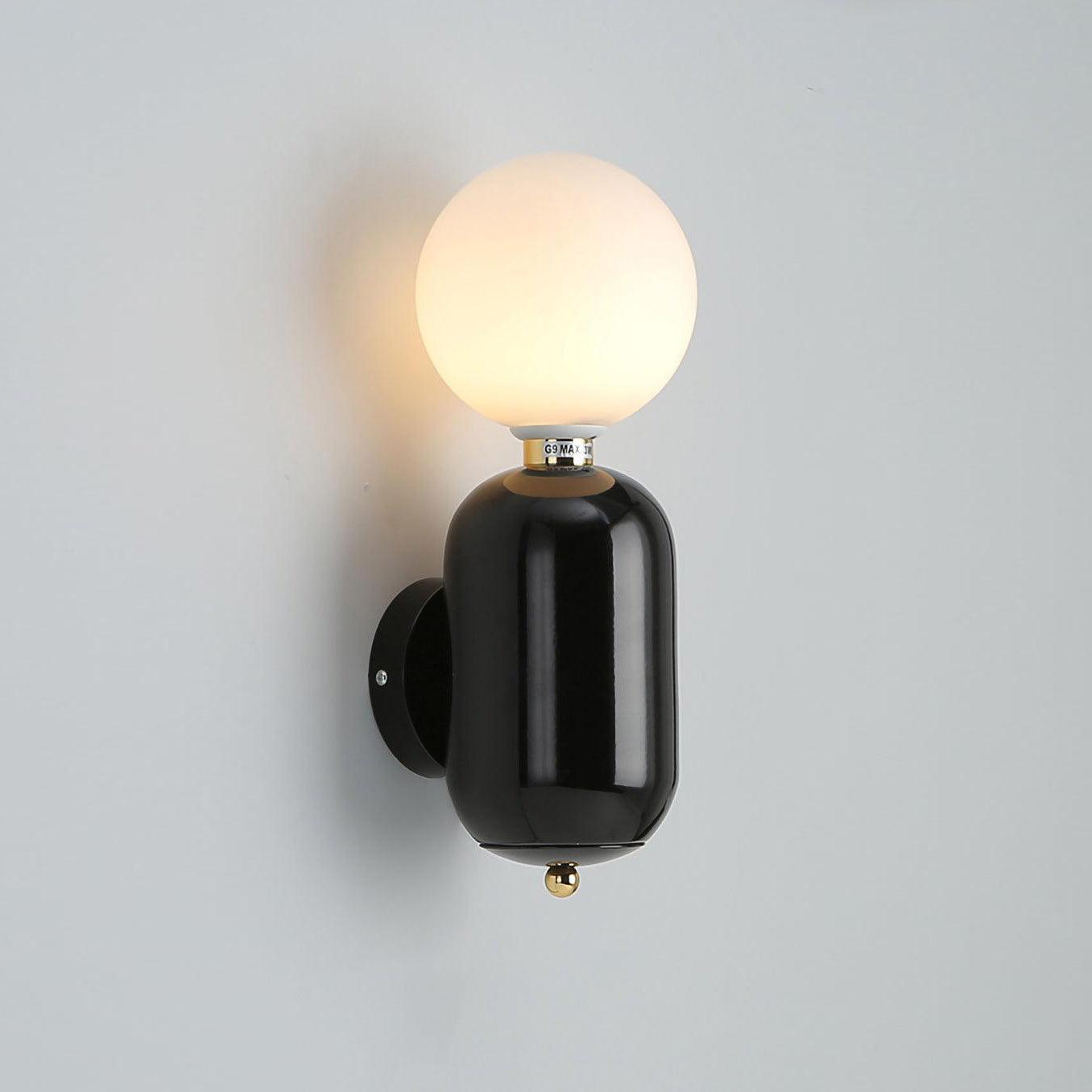 Black rounded wall sconce lit on gray wall, white globe, circular plate behind left and small lower gold-tone finial