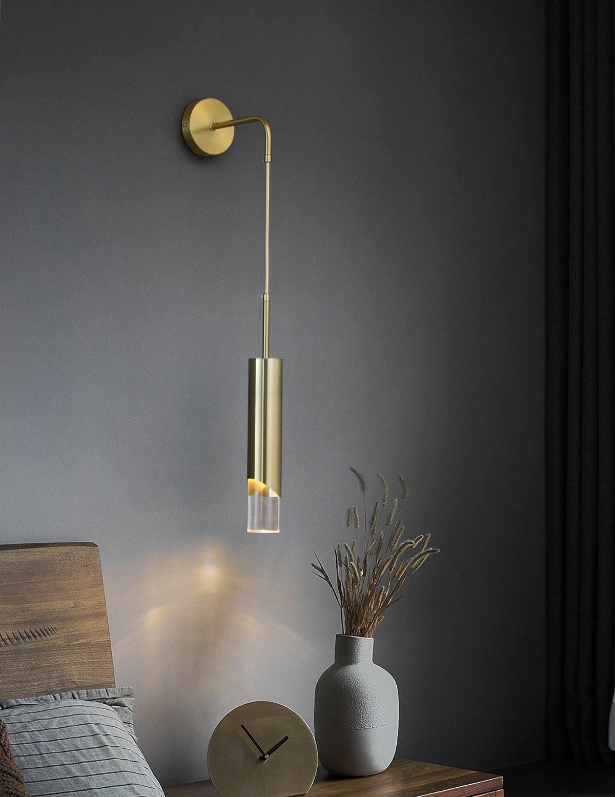 Gold wall light above bedside surface with round gold clock and two-tone gray vase, wooden headboard and dark curtain