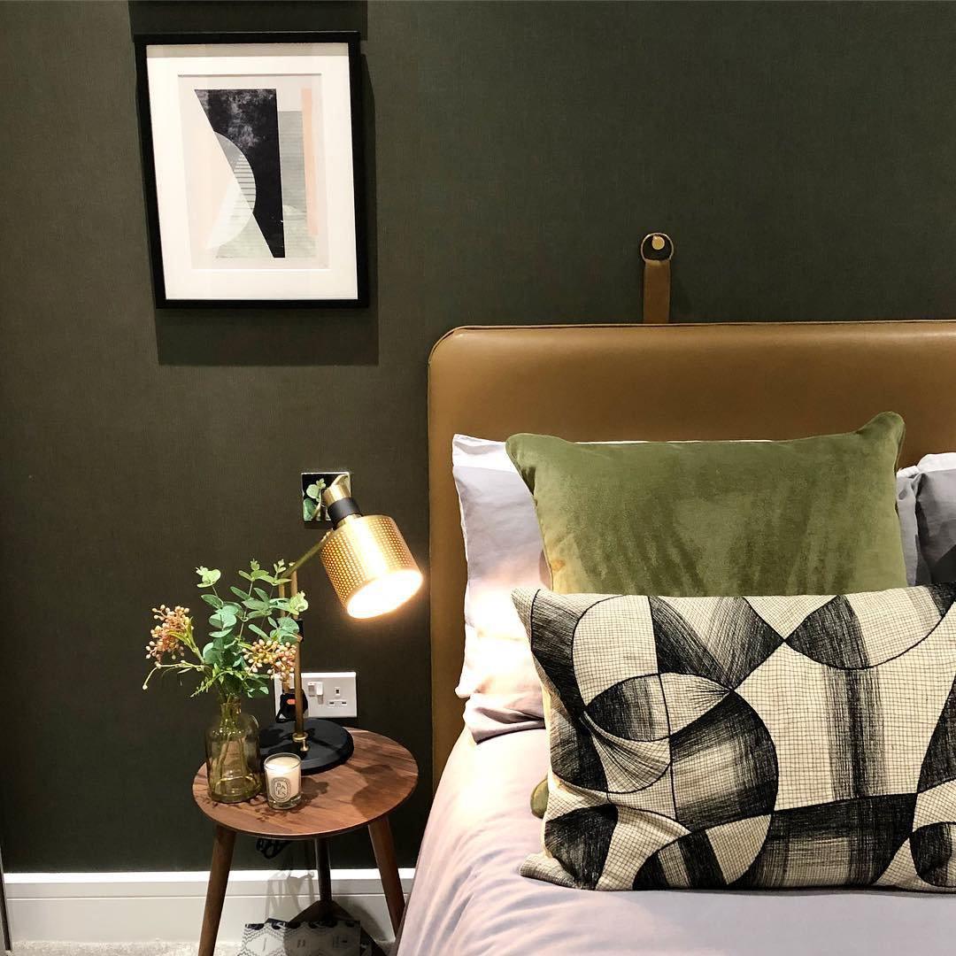 Lit gold-and-black single-head table lamp on a round bedside table beside flowers and a candle, with green bedding behind