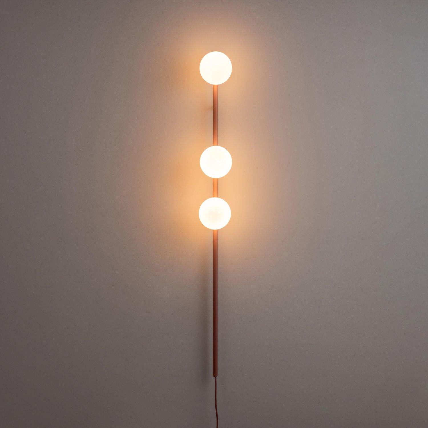 Pink three-globe wall light in a dim room, warm orange halos around illuminated white globes and long lower rod
