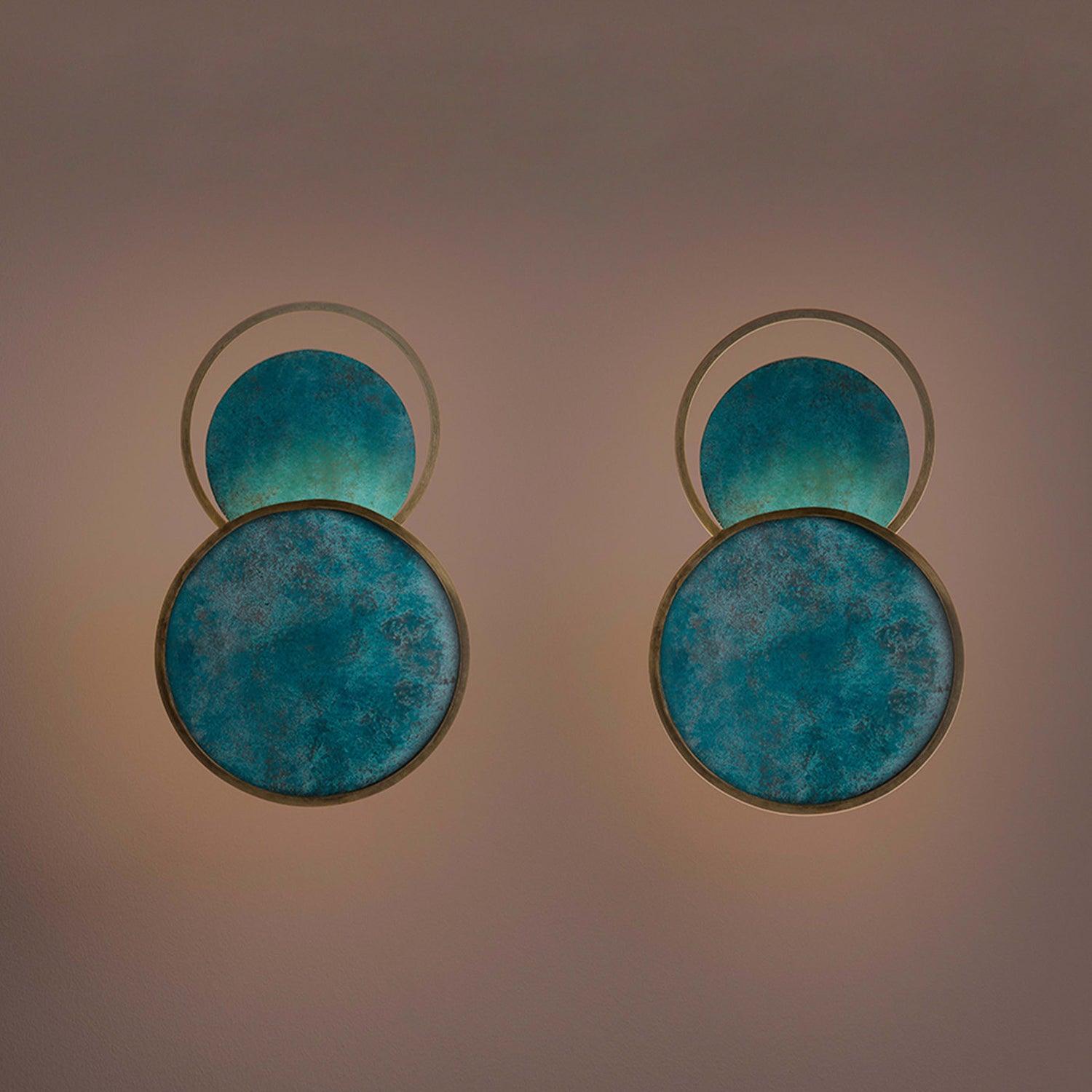 Two blue circular wall lights in a darker wall view, with overlapping faces, open upper rings and surrounding side light.