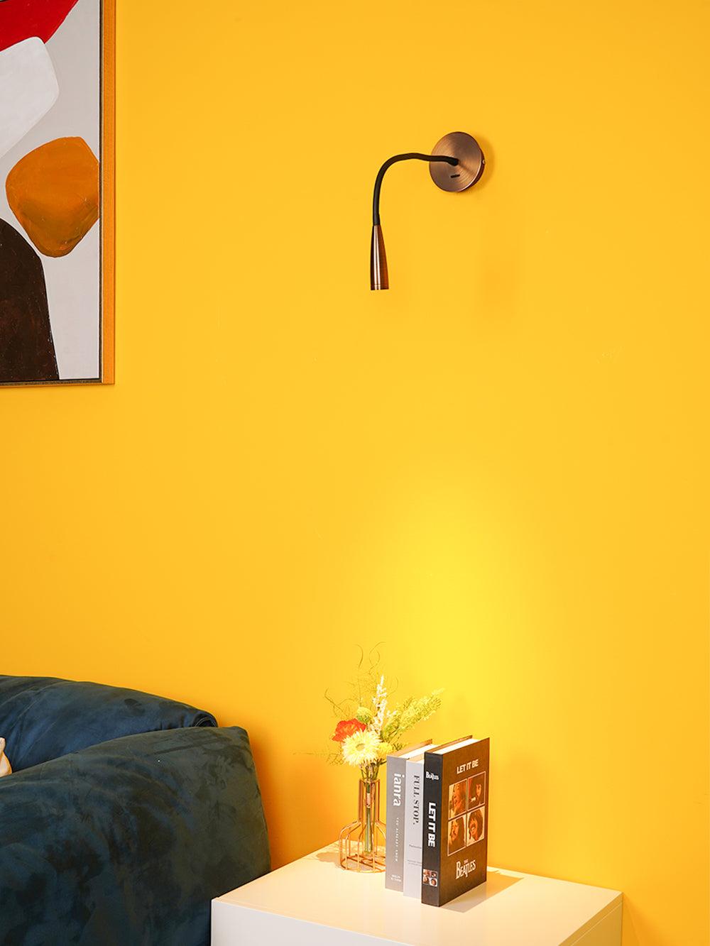 Close bronze reading light with sideways-curved black arm on yellow wall above cabinet, books and flowers