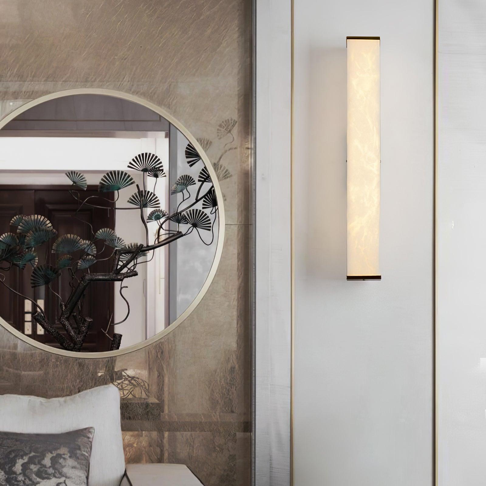 Front view of a glowing alabaster wall light with brass ends beside a round decorative mirror and pale sofa