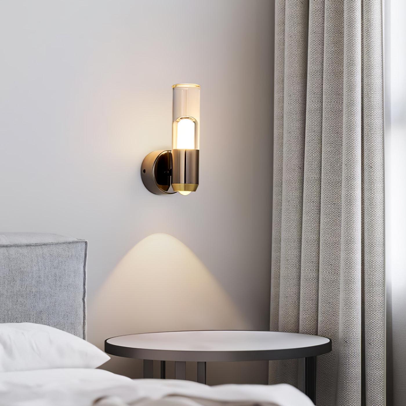 Illuminated clear-cylinder wall light above a round bedside table beside a grey upholstered bed and curtain