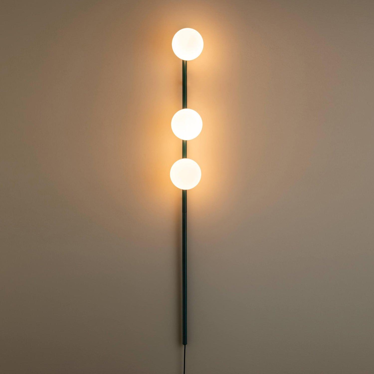 Dark teal three-globe light in a dim room, strong warm halos on wall and dark cable below vertical stem