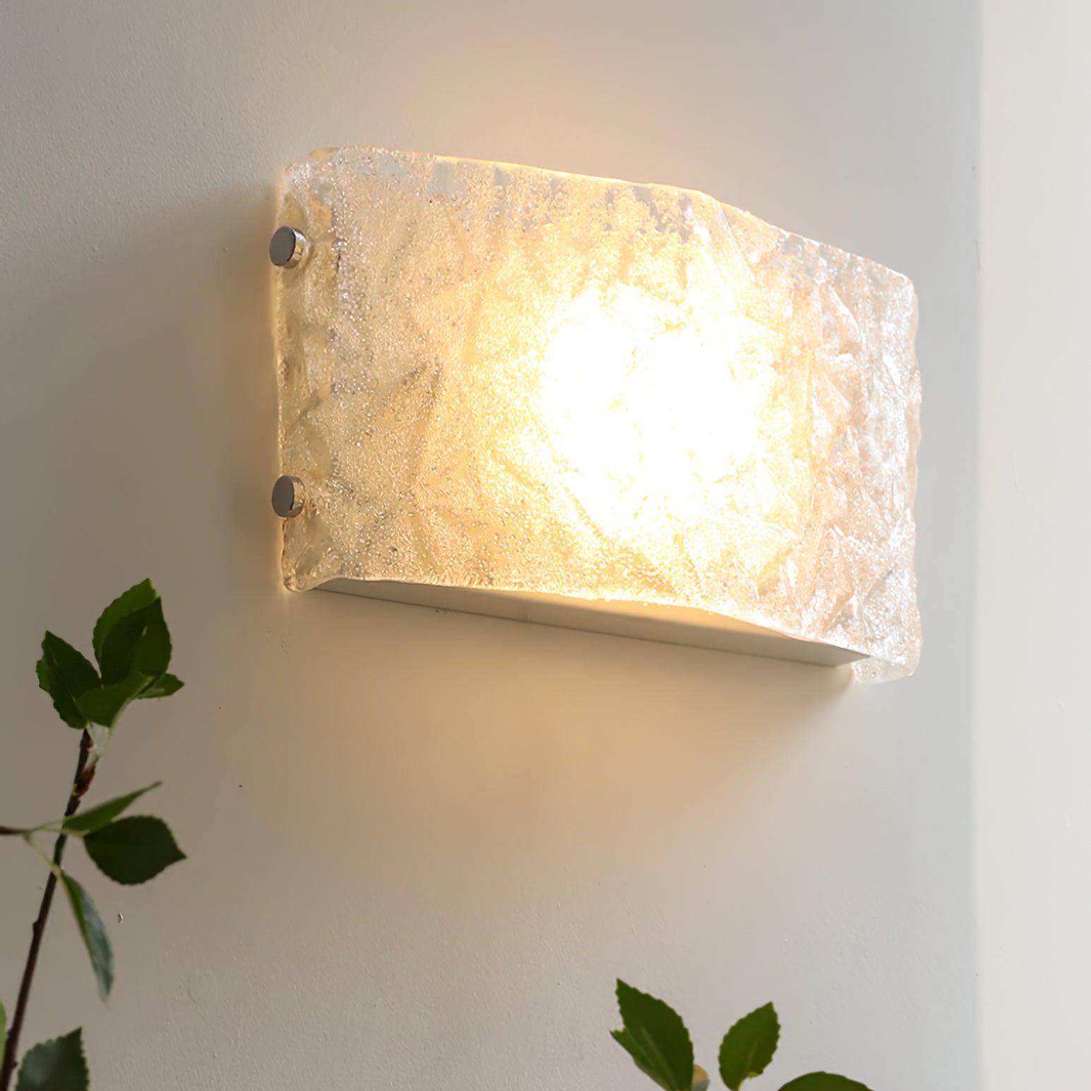 Angled illuminated glass wall light with warm-looking central glow, chrome fasteners and a leafy plant at the left