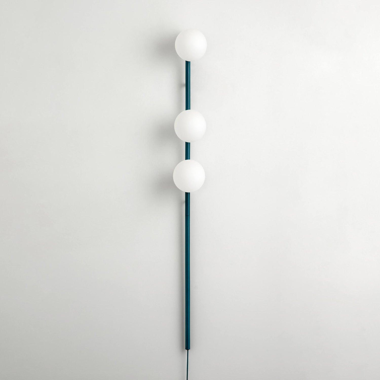 Unlit teal-stem three-globe wall light on pale wall, white glass spheres and dark cable below long lower rod