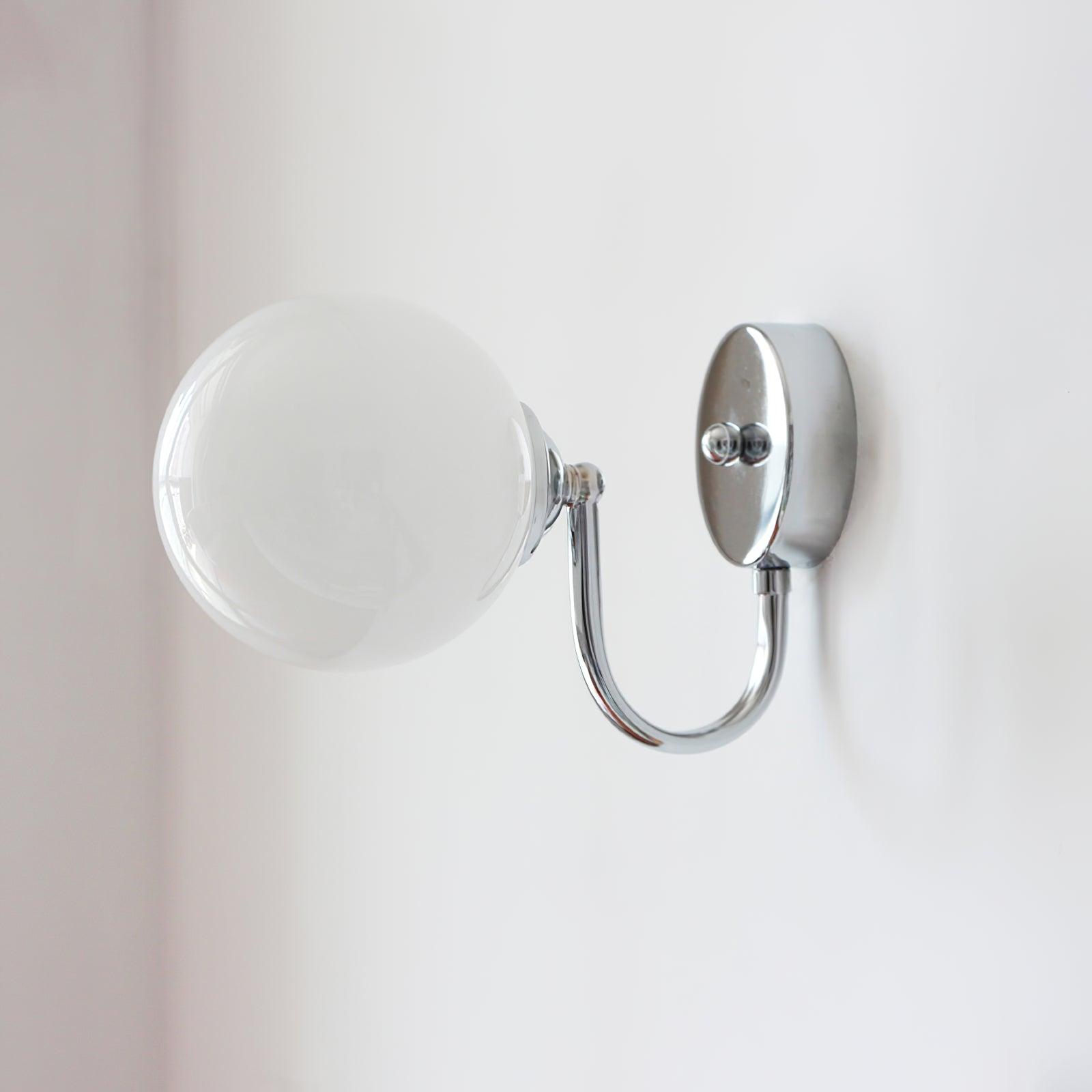 Close view of white globe, round chrome-coloured wall plate and lower curved arm against a pale wall