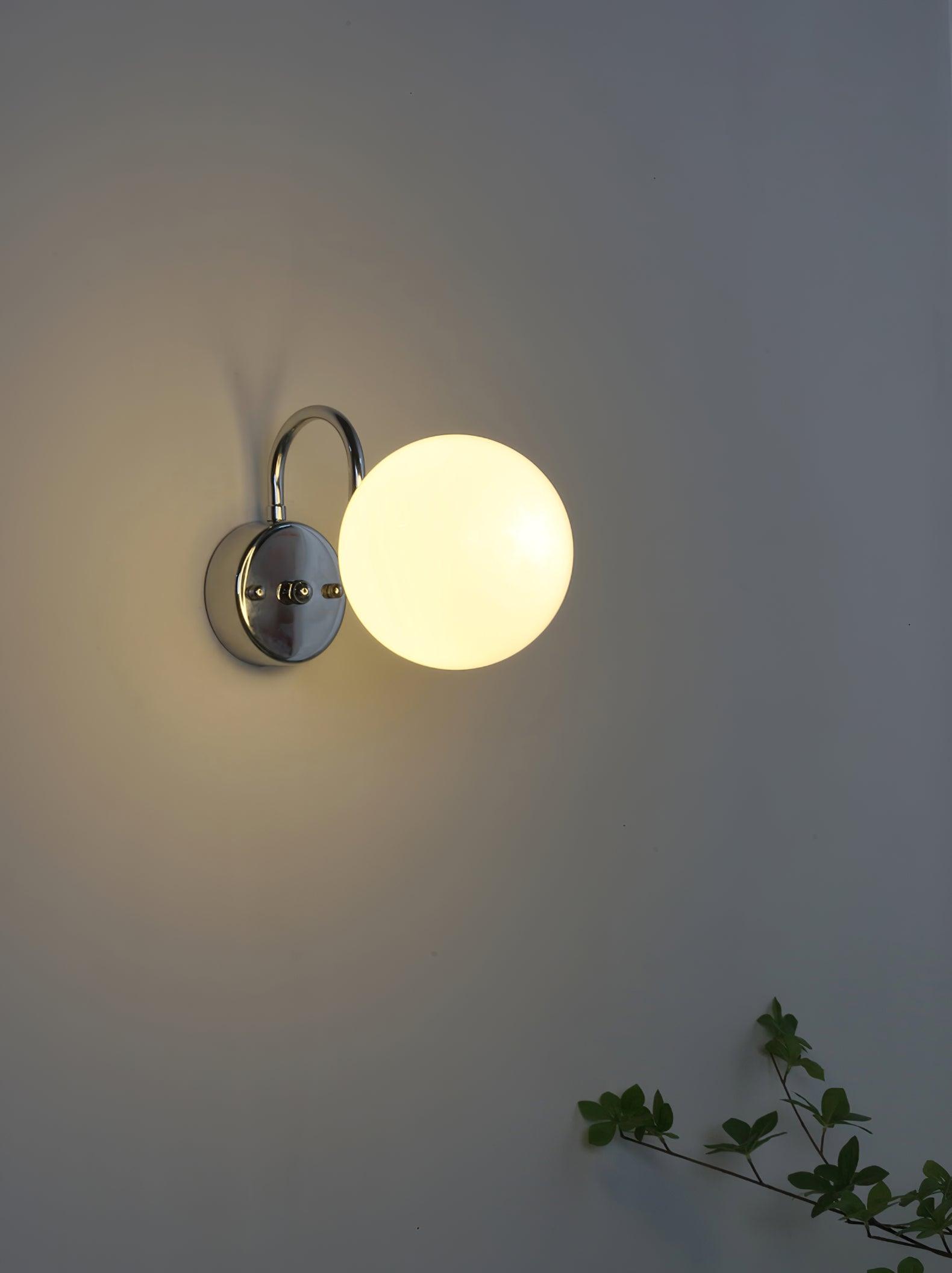 Glowing white globe at right of a chrome-coloured round plate, with arched arm above and foliage below on a pale wall