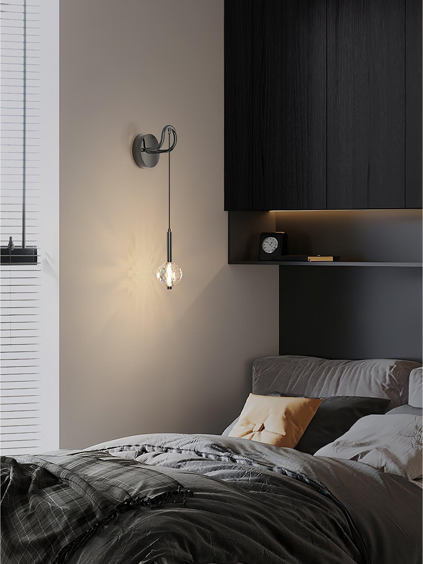 Hanging glass wall light beside dark bedding and overhead cabinets, with window blinds and clock on a recessed shelf