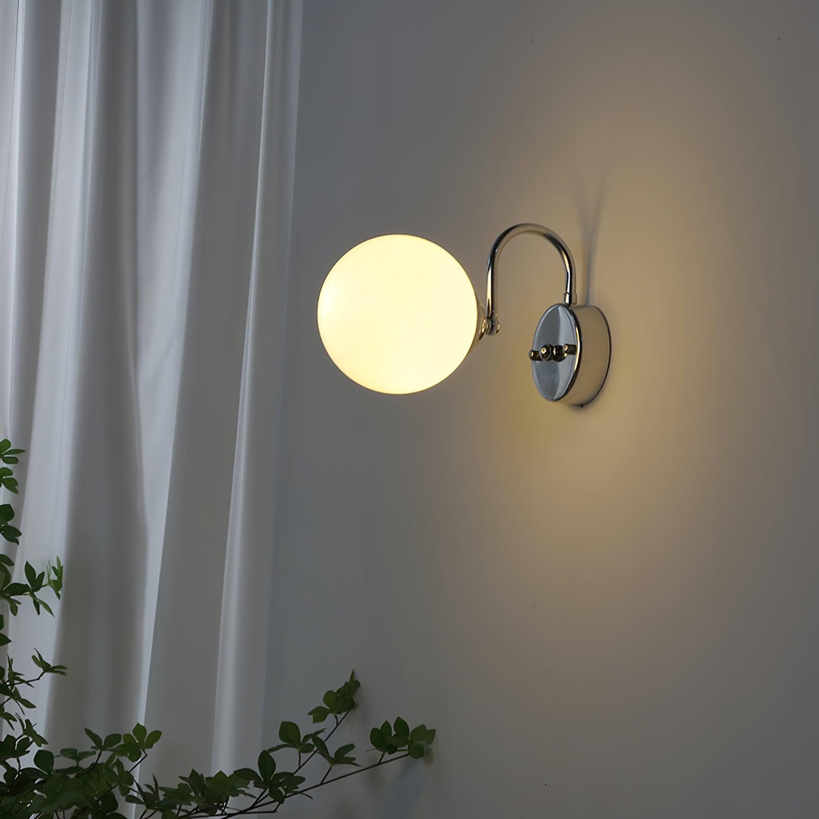 Lit Staggered globe at left of a round plate, with chrome-coloured arm arching above, beside a white curtain and green branches