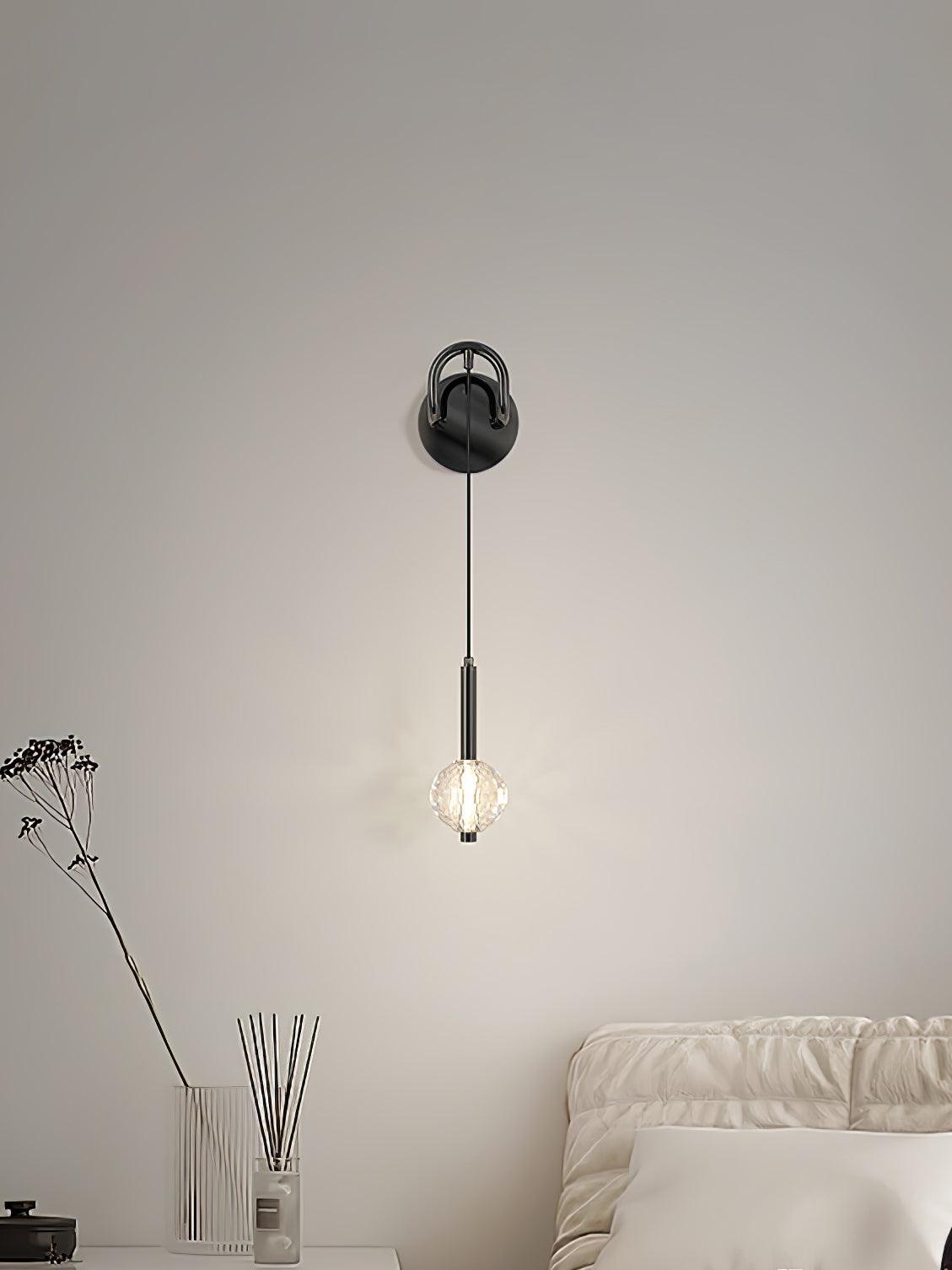 Closer view of lit faceted globe and long dark neck beside pale bedding, with stem vase and reed diffuser at left