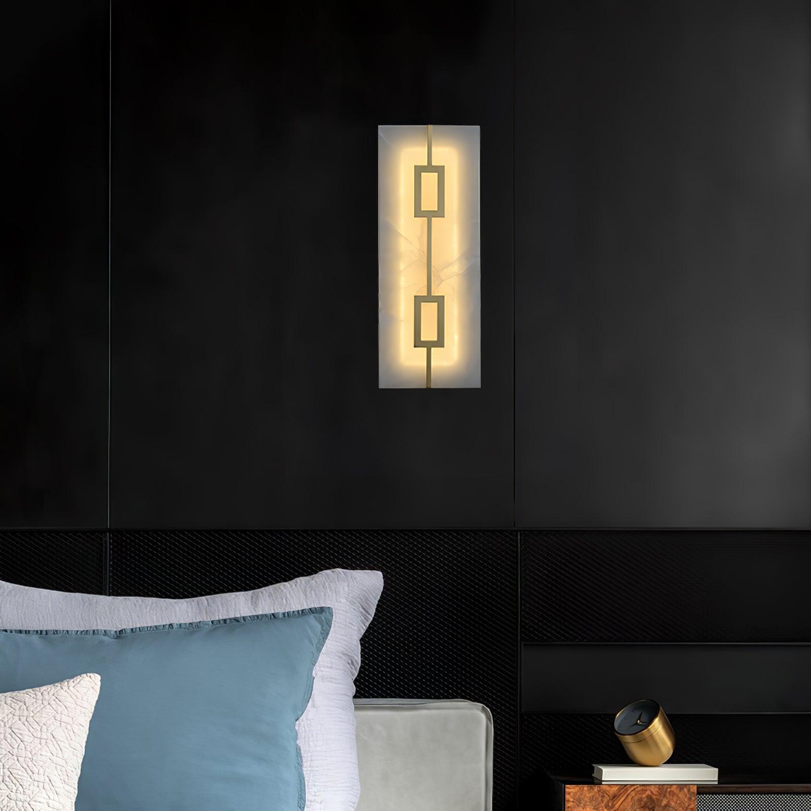 Tall geometric wall light on a dark bedroom wall above blue and white pillows, beside a cabinet and clock