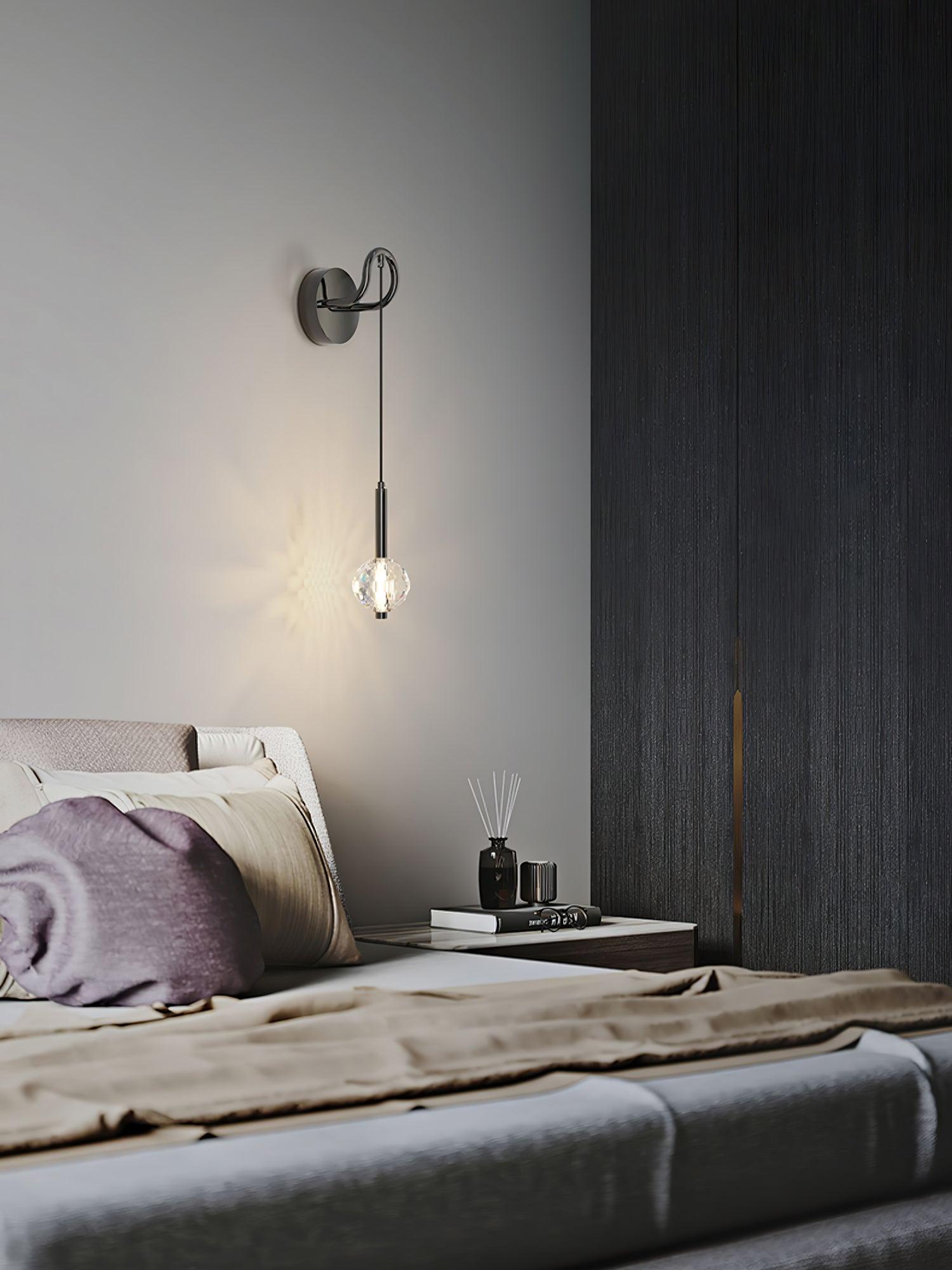 Lit suspended globe wall fixture beside grey headboard and dark wardrobe, above bedside books and reed diffuser