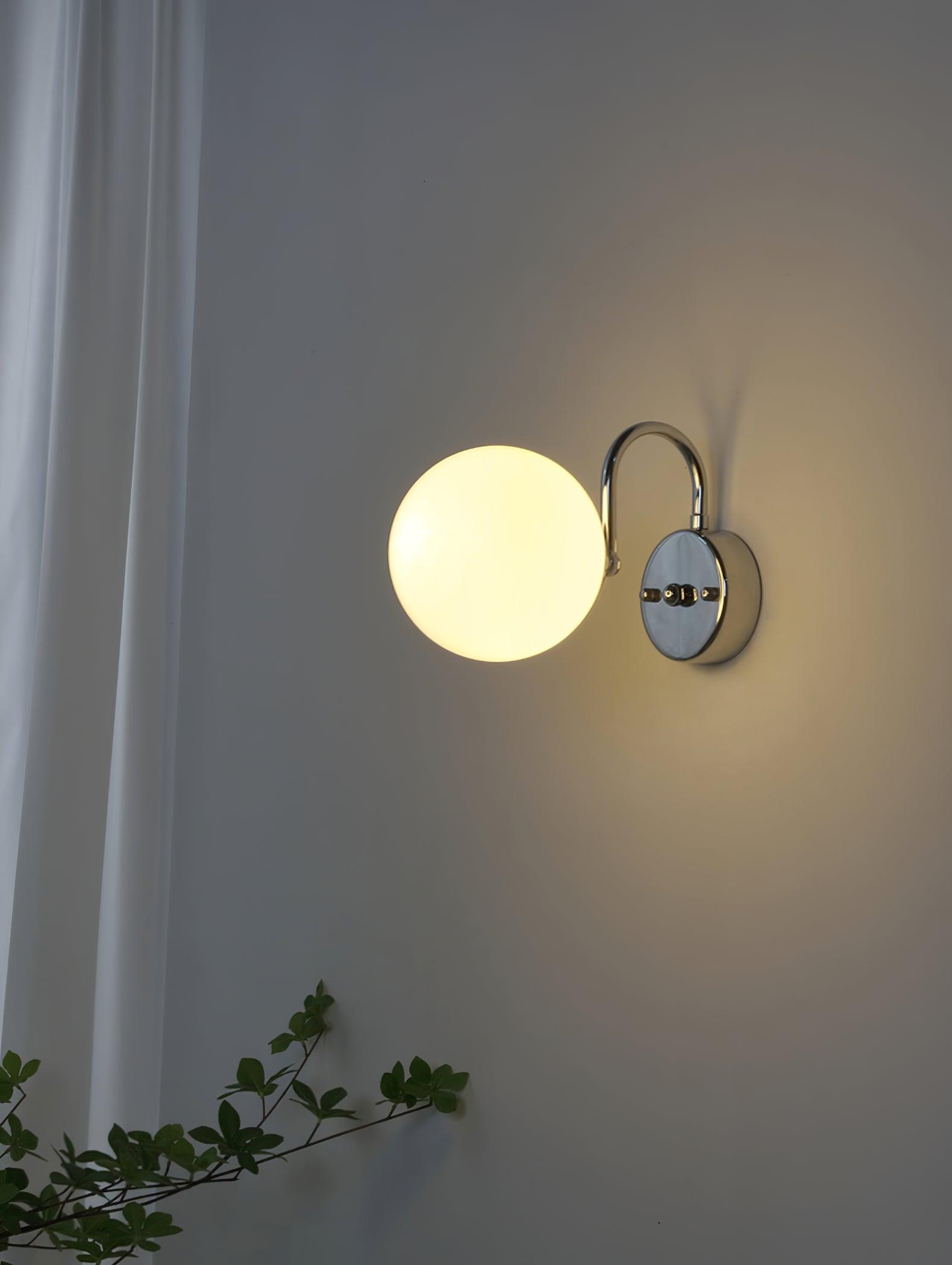 White globe glowing at left of a round plate with arched chrome-coloured arm above, beside a curtain and green branches