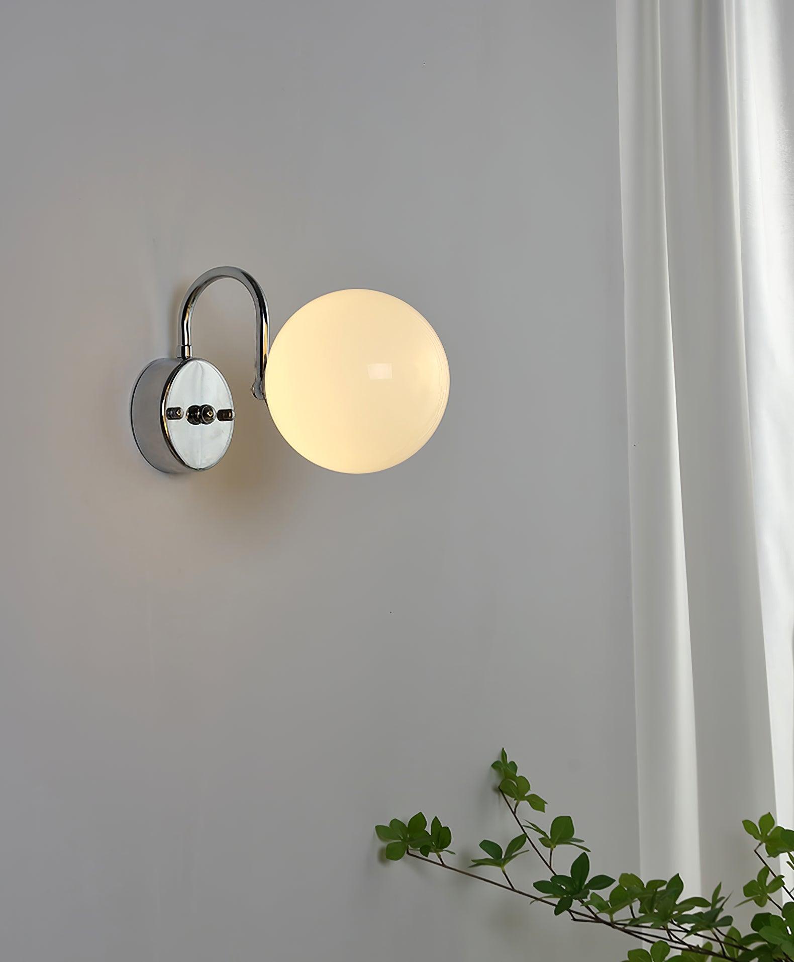 Lit globe wall light with plate at left and curved arm above, near a white curtain at right and leafy branches below