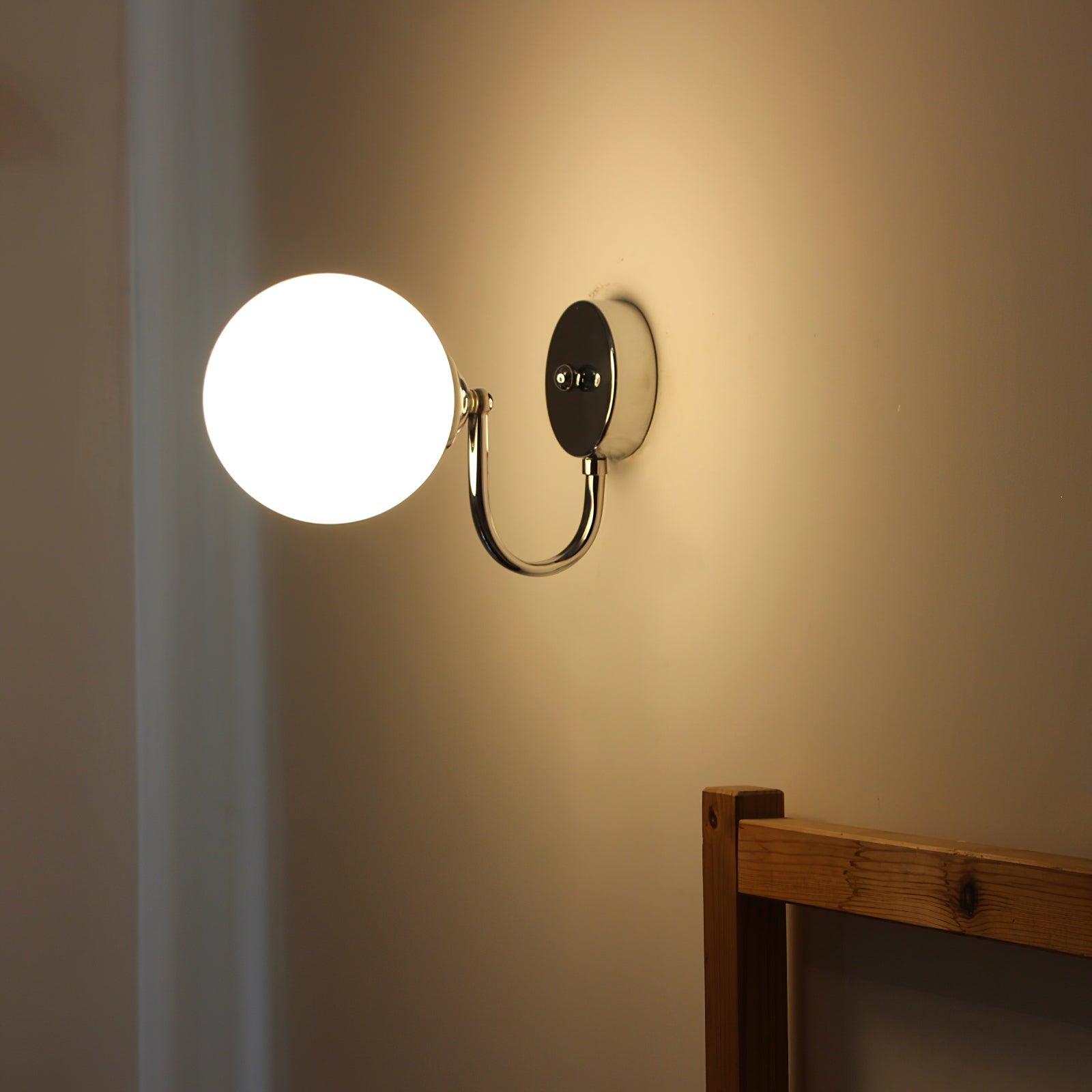 Lit white glass globe with chrome-coloured plate and lower curved arm on a cream wall above a wooden headboard