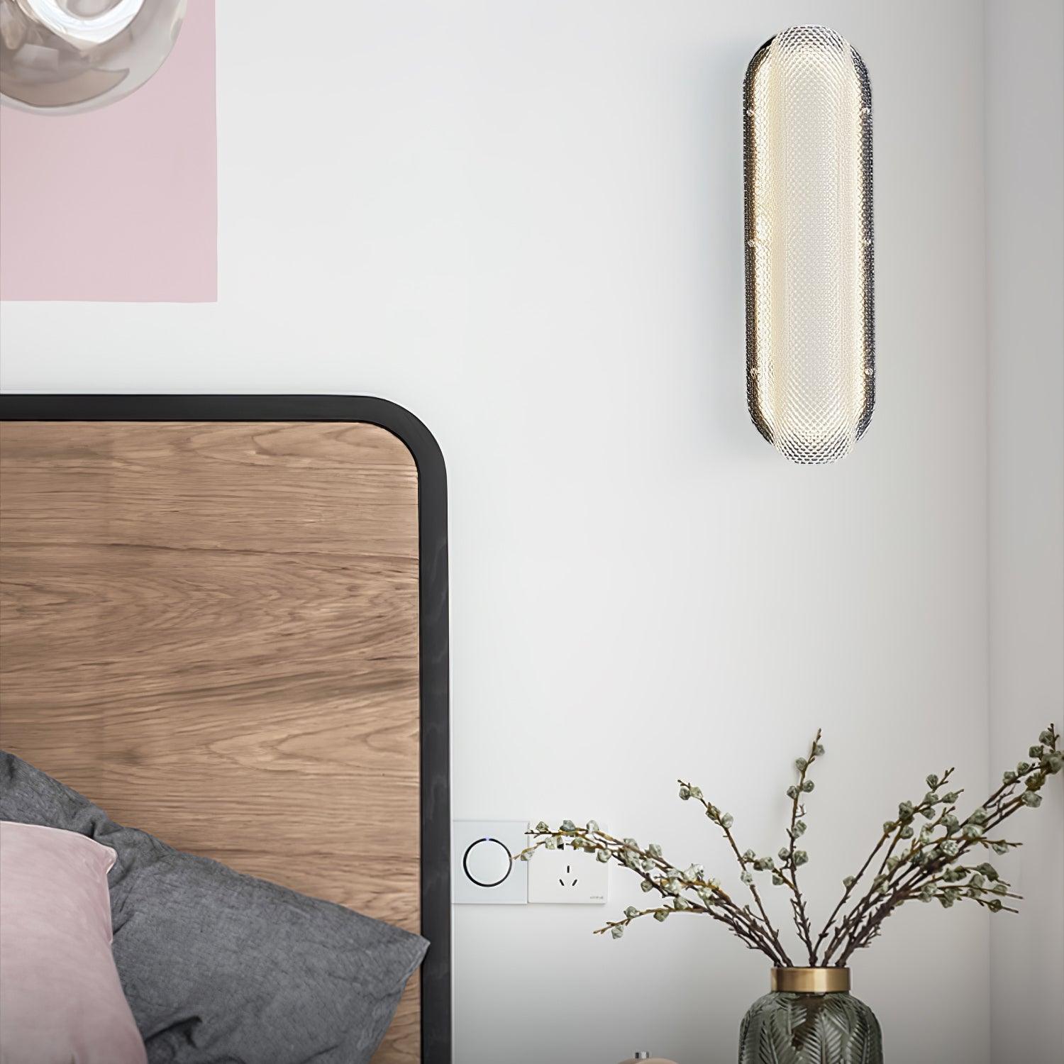 Vertical Vale glass wall light beside a wooden bed headboard and a vase of flowering branches