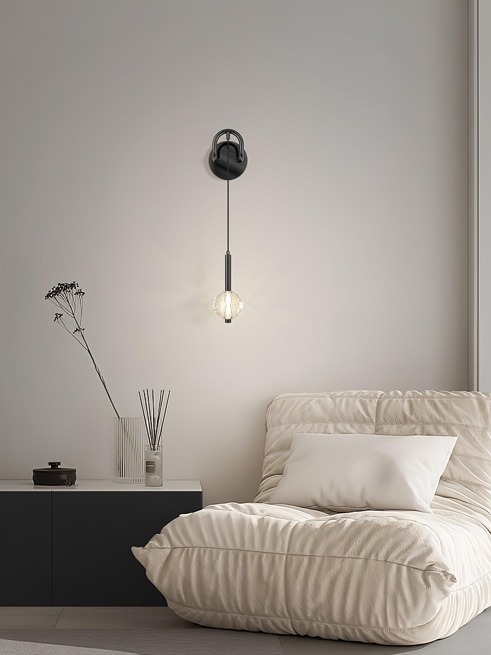 Wide bedroom view of a lit hanging-globe wall light above pale bedding and dark bedside chest with stem vase