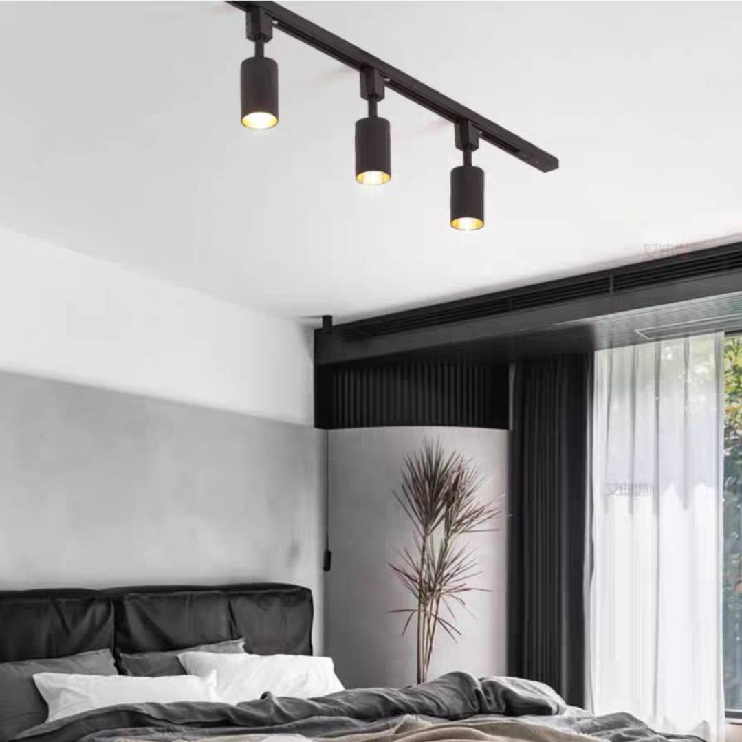 Black Freja three-head track above a dark padded bed, pale pillows, black curtains and a slender potted tree.