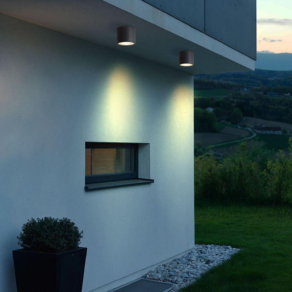 Two black Orr ceiling cylinders beneath an exterior overhang above a small window, beside a planter, gravel strip and lawn.