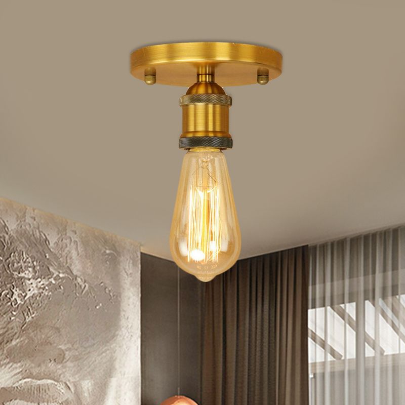 Bright brass-tone Alessio canopy and knurled short holder above an elongated glowing bulb, beside a textured wall and curtains.