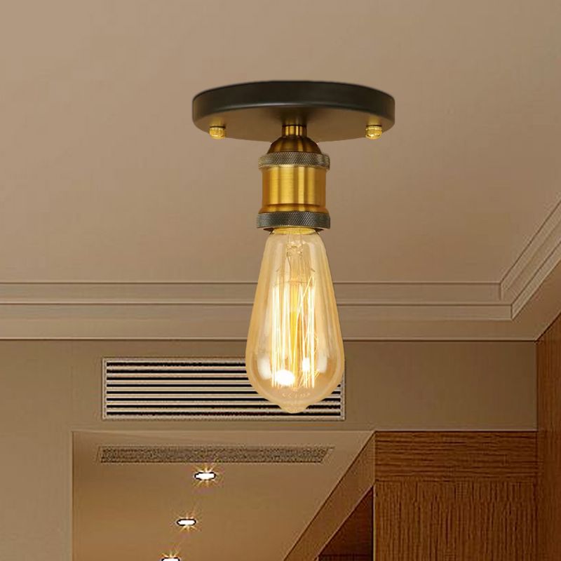 Close brass-tone Alessio holder and darker round canopy above an elongated glowing bulb, with a vent grille and wood wardrobe behind.