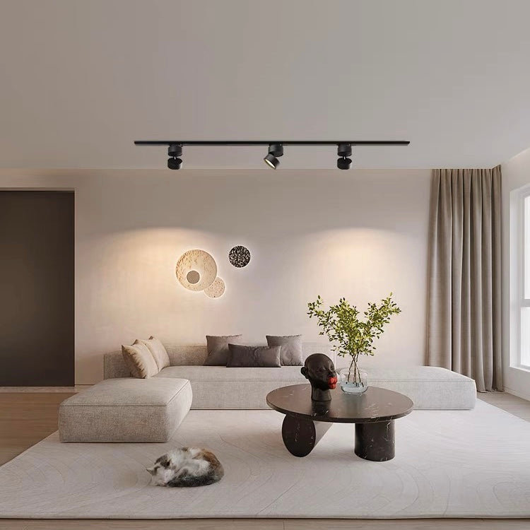 Black cylindrical heads along a rail above a light grey sectional sofa, black round table, plants and wall ornaments.