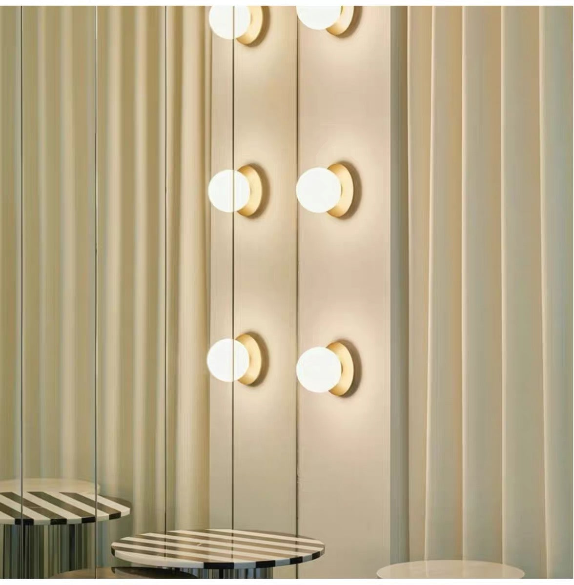 Gold Valentina smooth-globe lights along a mirror, with reflections, beige curtains and a round striped stool.