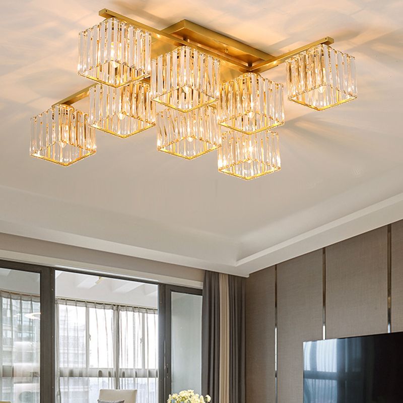 Wider room view of eight gold-toned Kristy cube shades above a window, curtains, wall panels, partial TV, seat and yellow flowers.