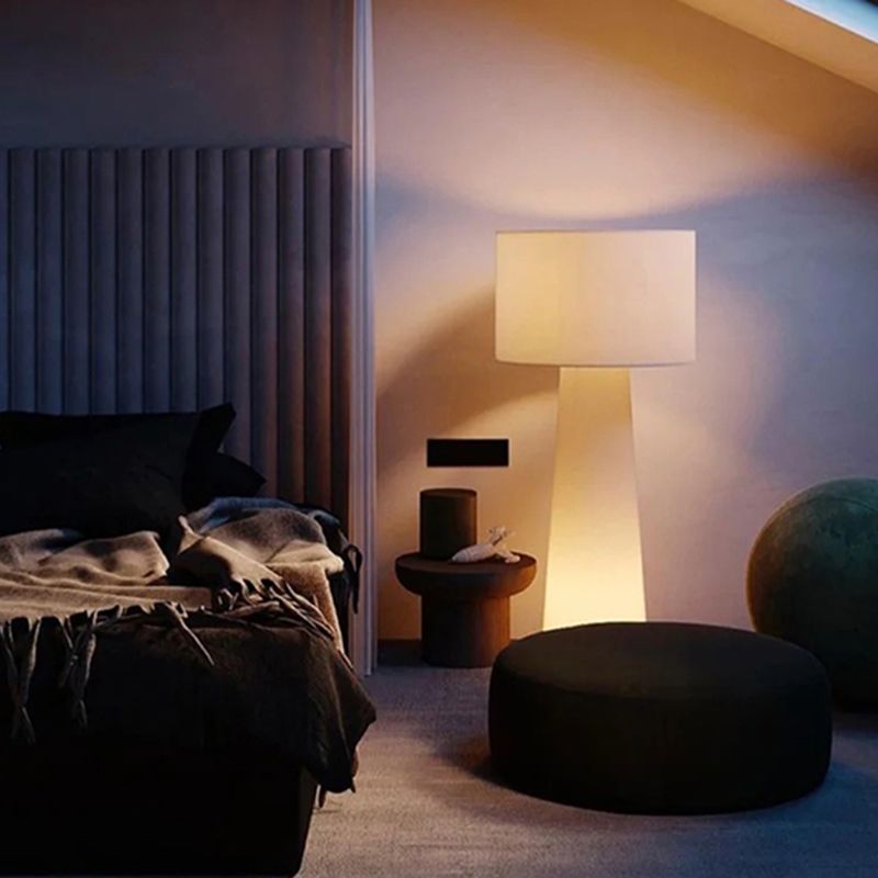 Glowing drum-shade floor lamp beside a dark bed, small round table and ottoman in a softly lit room