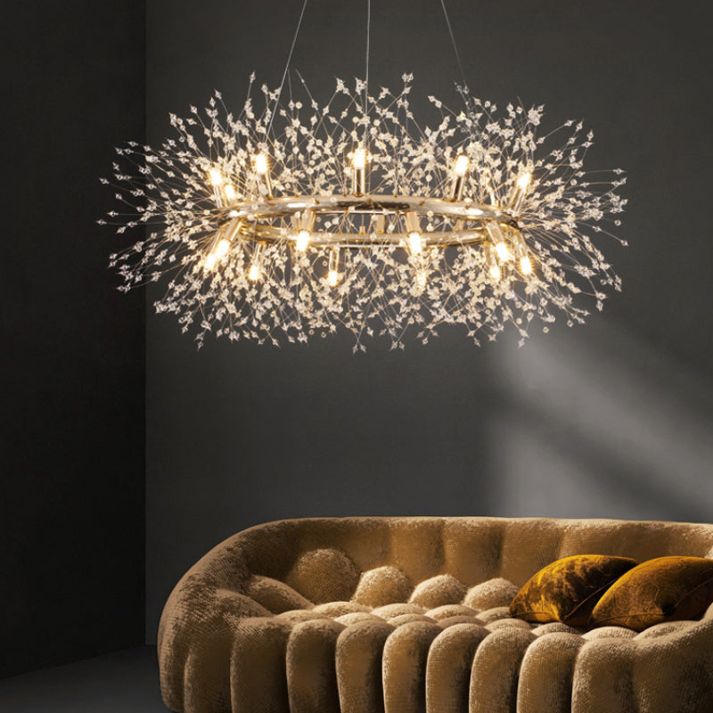Lili gold-coloured crystal ring pendant before a dark paneled wall, above a beige rounded tufted sofa with brown cushions.