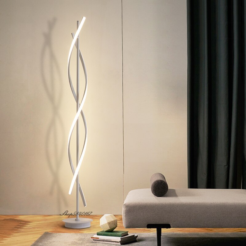 Illuminated white Louise lamp with crossing curved light bars and round base beside a gray chaise