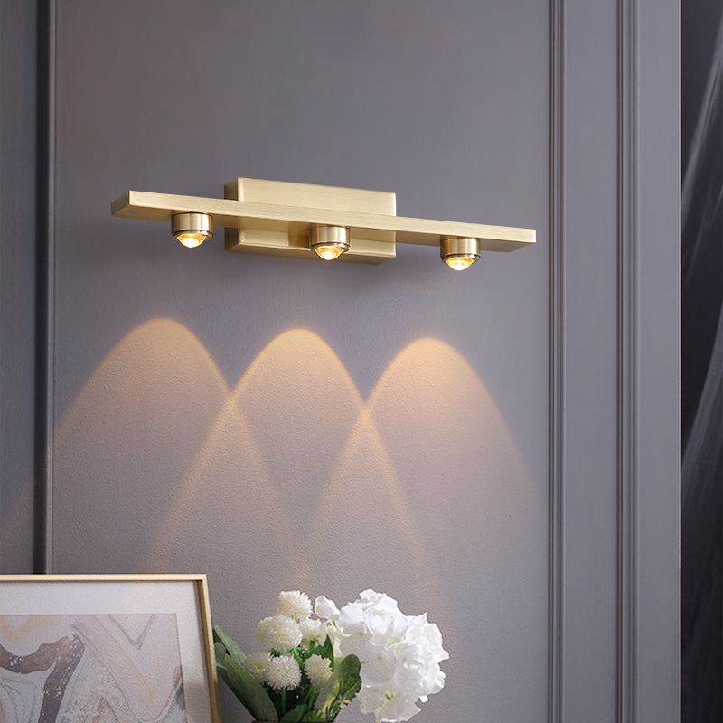 Angled Leigh three-light gold-tone wall lamp above a framed picture and flowers on a dark wall