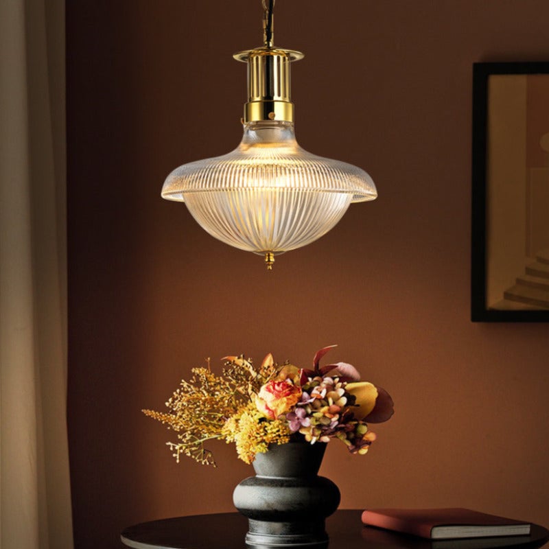 Alessio glass pendant glowing above a flower vase against a warm brown wall.