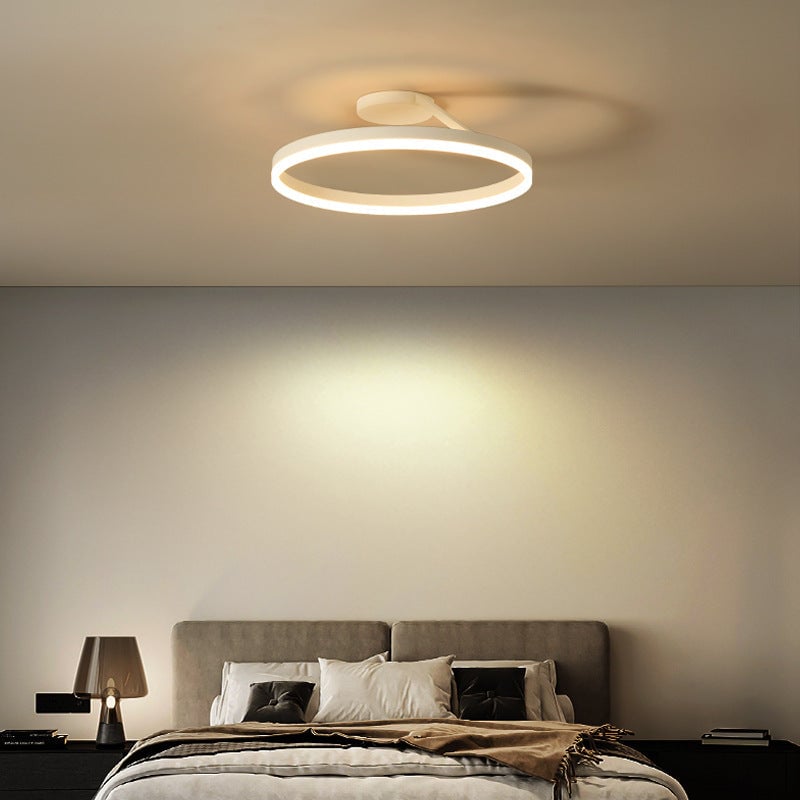 White illuminated ring ceiling fixture above a bed with gray headboard, pillows and a table lamp