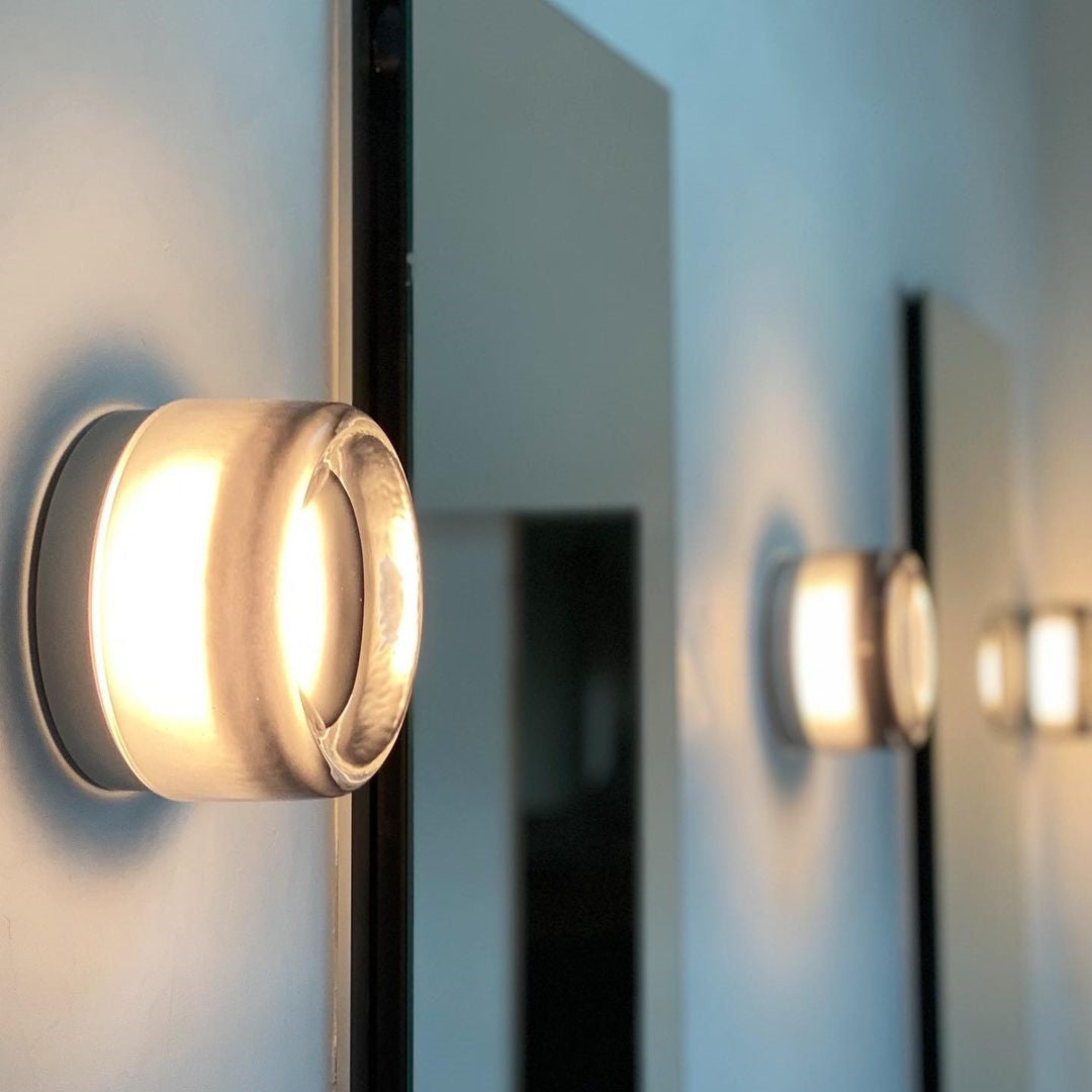 Close pale circular wall light with a luminous side band beside upright mirrors and another light in the distance