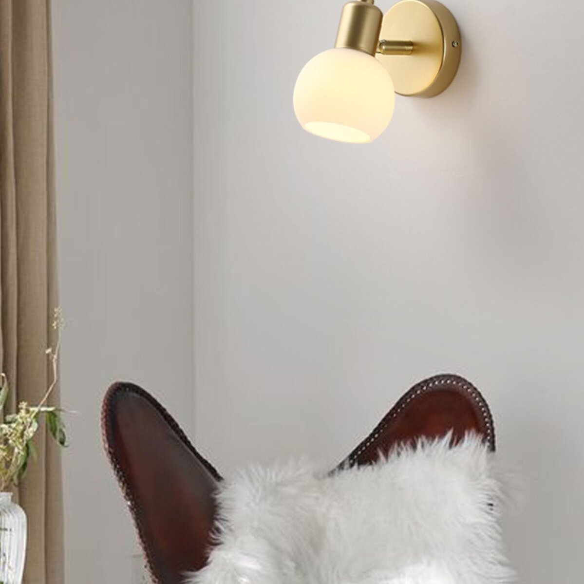 Single illuminated Gold globe wall light above a brown wing chair with a white furry cushion beside a window