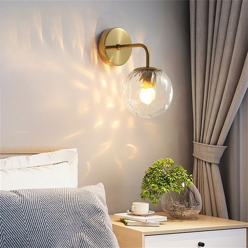 Gold Valentina glass globe wall light above a bedside table with books and cup, beside curtains