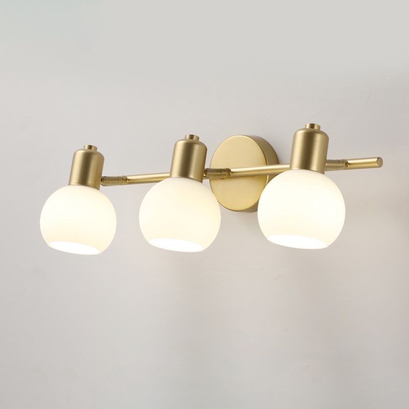 Three illuminated white globe shades on a Gold bar and round wall plate, viewed against a pale wall