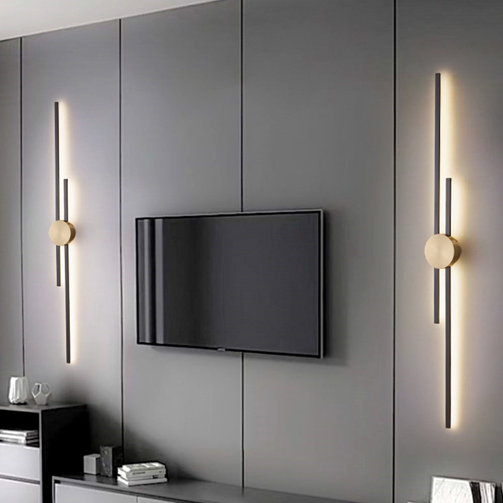 Two separate dark two-bar wall lights with gold-coloured discs flanking a television on a grey panelled wall