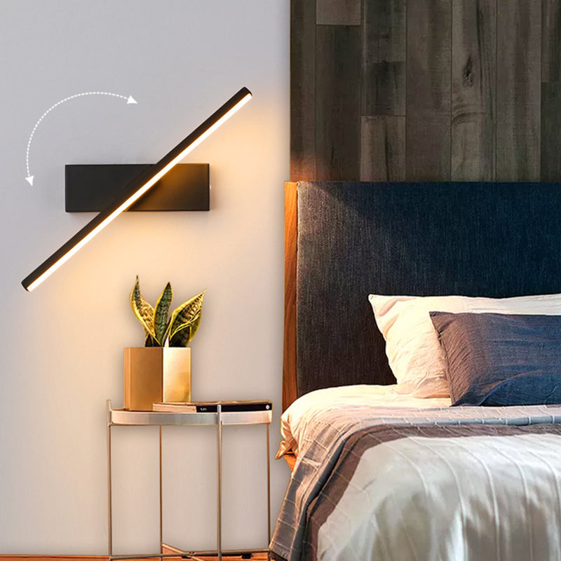 Illuminated black bar wall light beside a bed with a curved rotation arrow